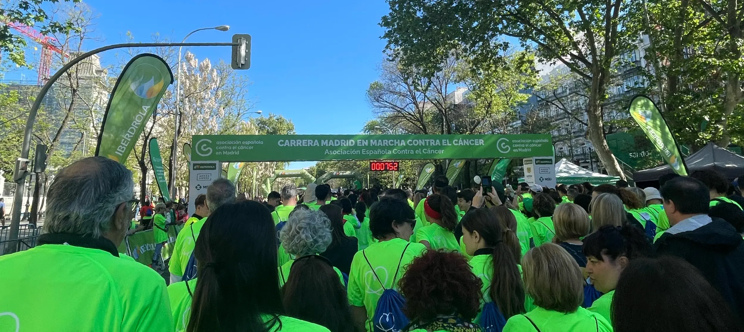 Participants in vibrant green shirts stand at the starting line of the "Carrera Madrid en Marcha Contra el Cáncer" race. The event's banner, sponsor logos, and a timer reading "00:07:52" are prominently displayed under a clear blue sky with surrounding trees and buildings.