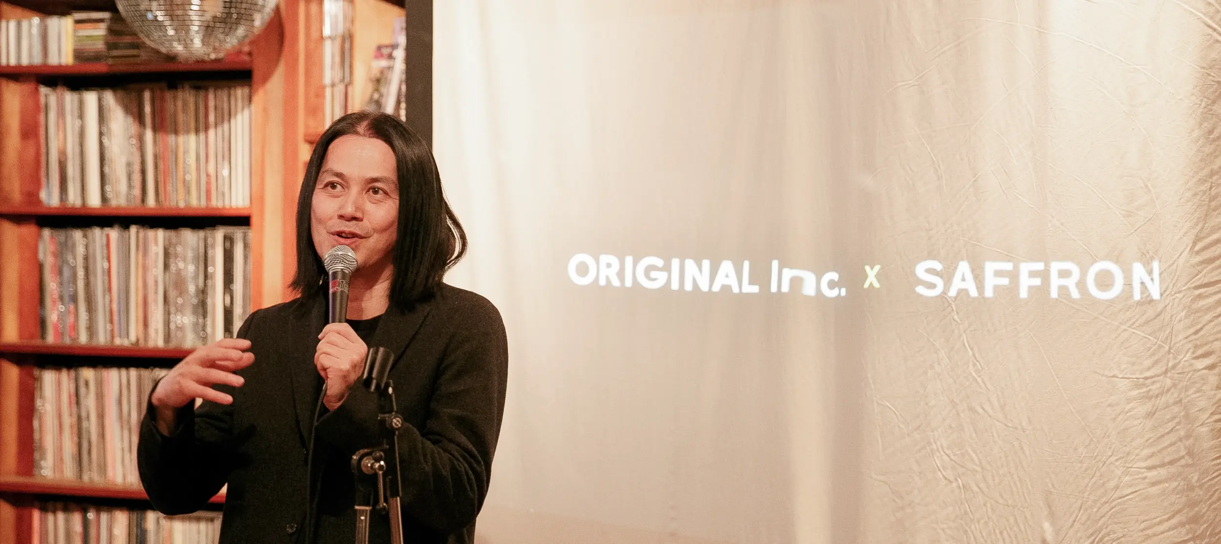 A speaker at the Saffron and Original Inc. event in Tokyo holds a microphone, addressing the audience. Behind him, a projected screen displays the event title "ORIGINAL Inc. x SAFFRON." The event is set in a room with shelves filled with records.